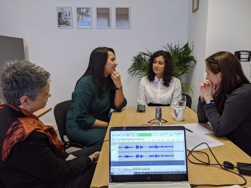 Sitting around a desk: Nicola, Kiran, Amna and Vicky. There is a computer on the foreground and a microphone on the desk