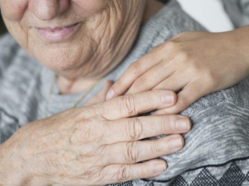 close up of a shoulder and face of an elderly person, their hand in holding someone else's hand which is over their shoulder