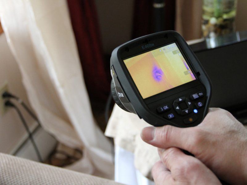 A person is holding a device that is pointing to a plug. the device's monitor reveals a purple colours around the plug, as it energy was 'leaking'